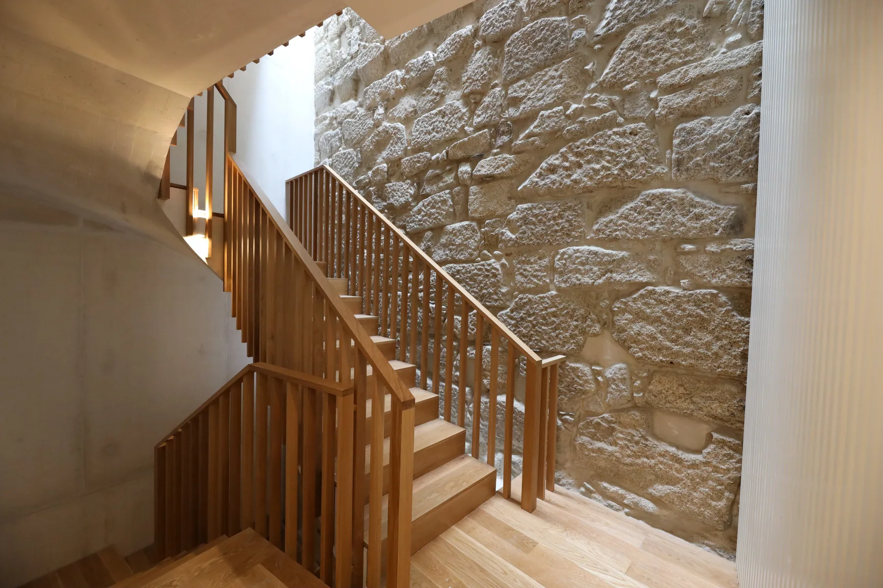 A wooden staircase rises beside an exposed stone wall in a bright interior.