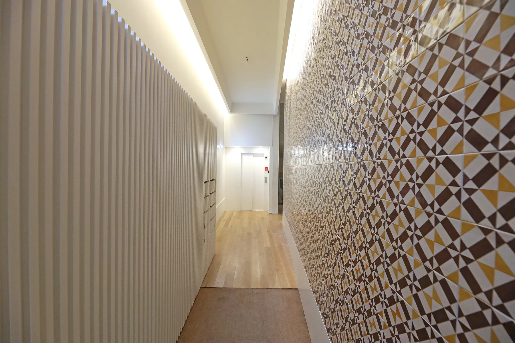 A narrow interior hallway has a patterned tile wall and light wood flooring.