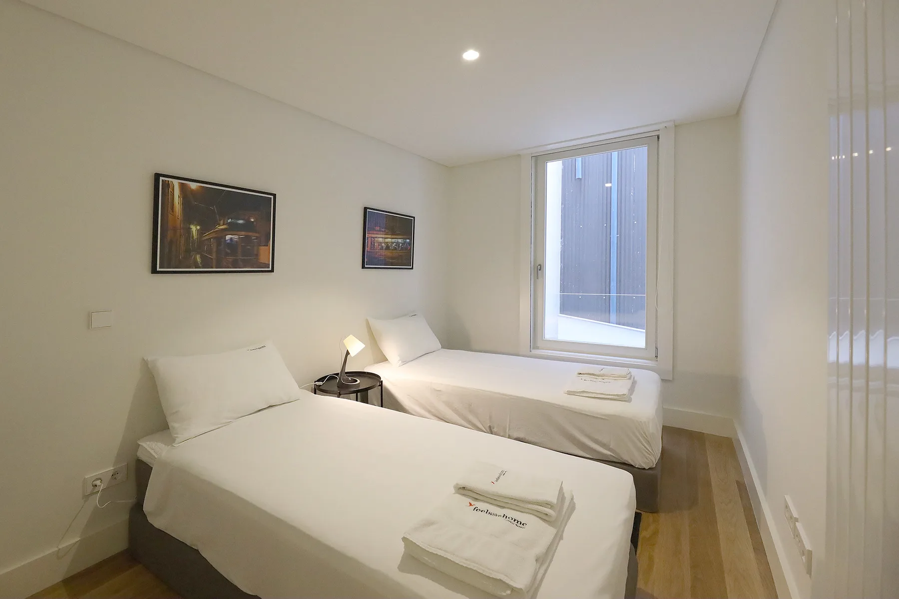 A bright bedroom with two single beds, framed pictures, and a large window.