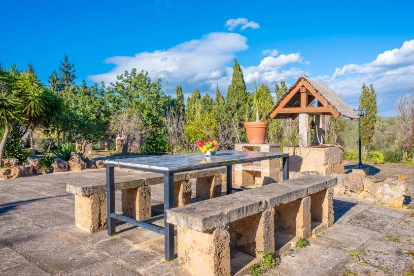 An outdoor stone dining area has a long table and benches among trees.