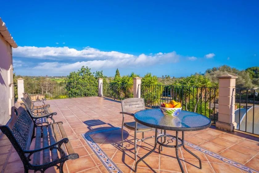 A sunlit outdoor terrace with a round table, chairs, and a railing overlooking greenery.
