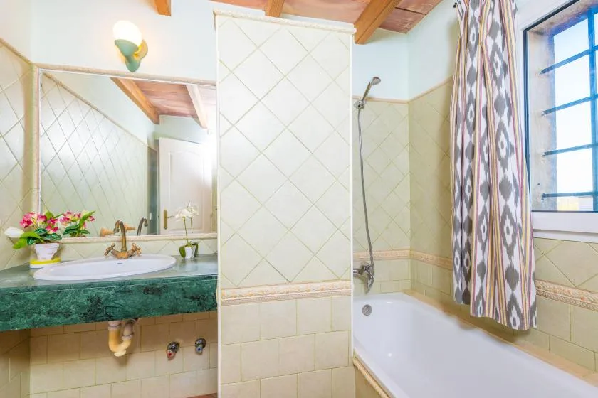A tiled bathroom with a sink, mirror, and bathtub beside a window.