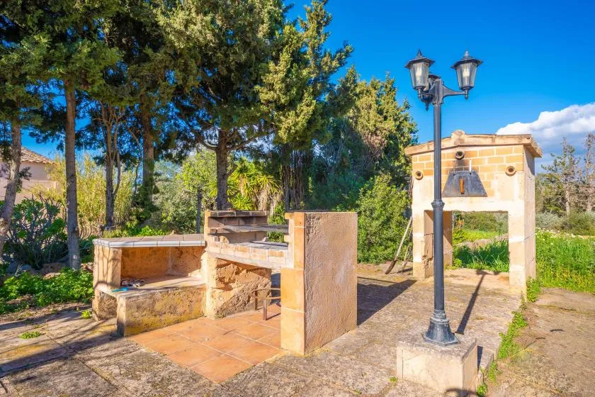 A stone outdoor barbecue area sits beside a garden entrance and tall trees.