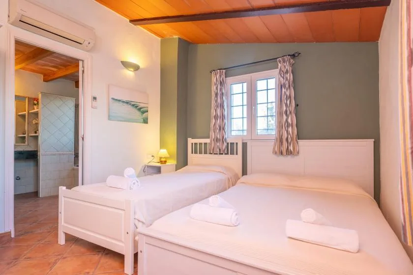 A bedroom with two beds, a window, and a warm-toned wooden ceiling.