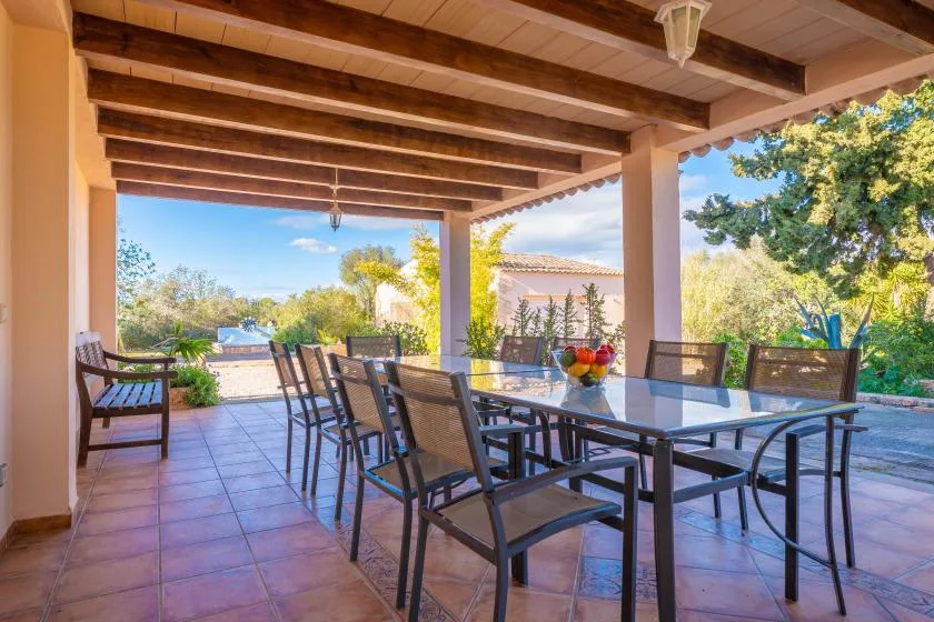 A covered patio has a long dining table and chairs, with trees visible beyond.