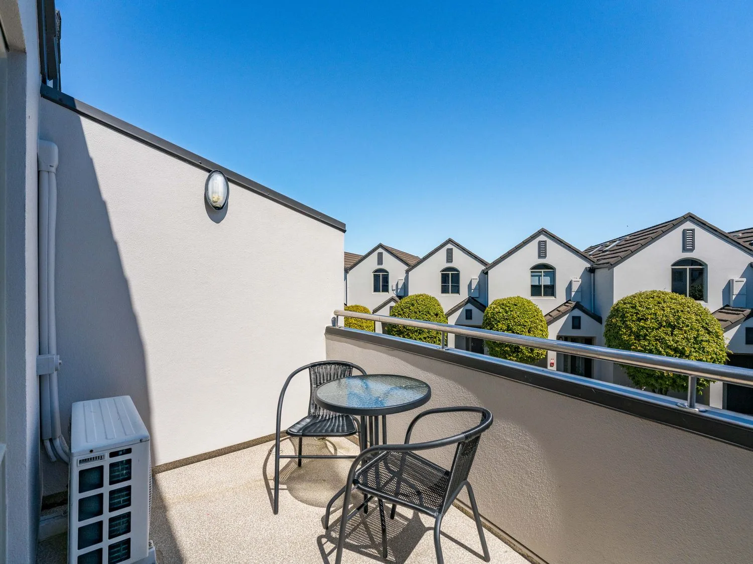 A balcony with two chairs and a round glass table overlooking houses with trimmed trees at Sun and Swim - Lake Taupo Holiday Home in Taupo Central
