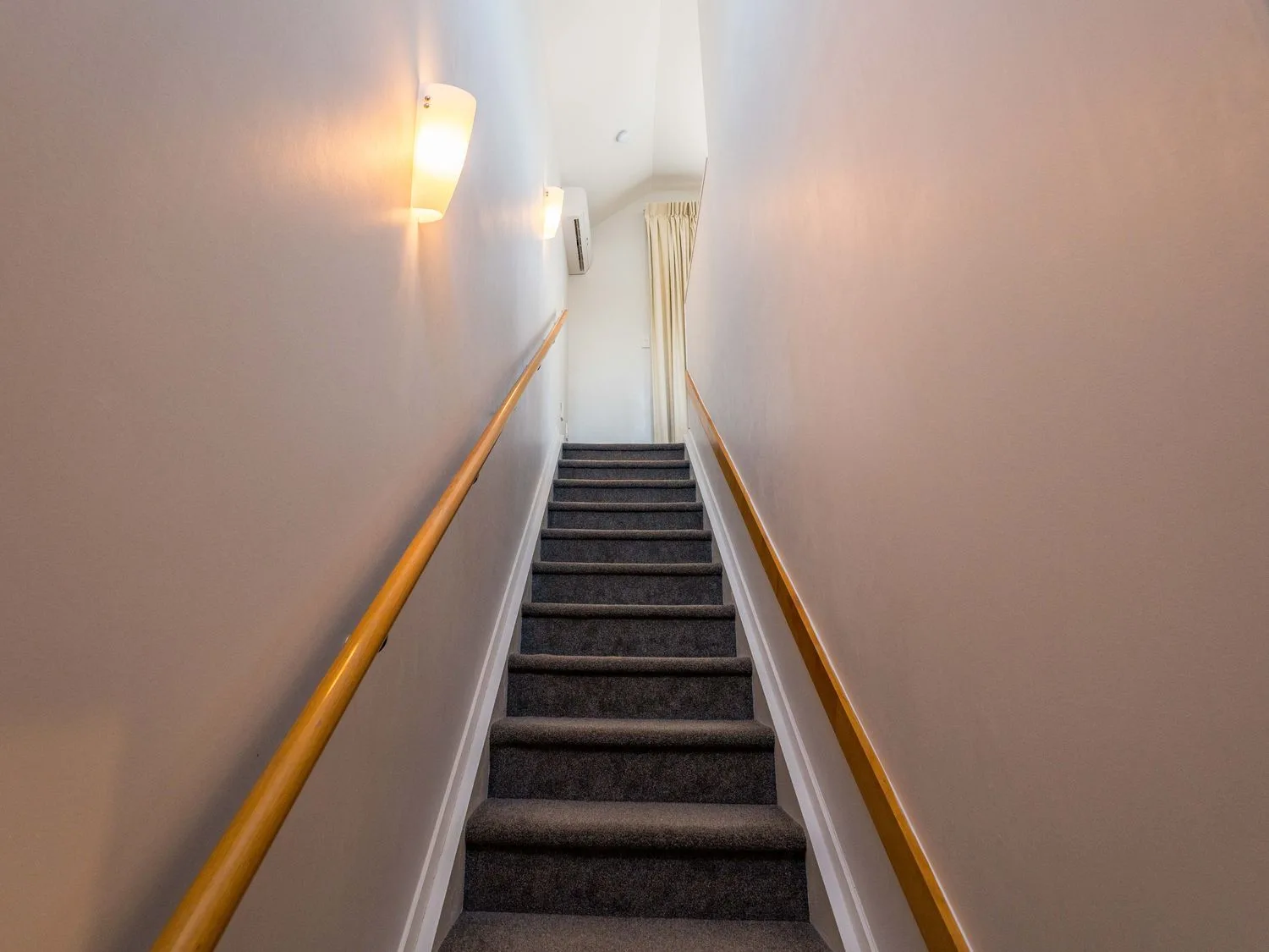 A carpeted staircase with wooden handrails and wall lights leading to a room with curtains at Sun and Swim - Lake Taupo Holiday Home in Taupo Central