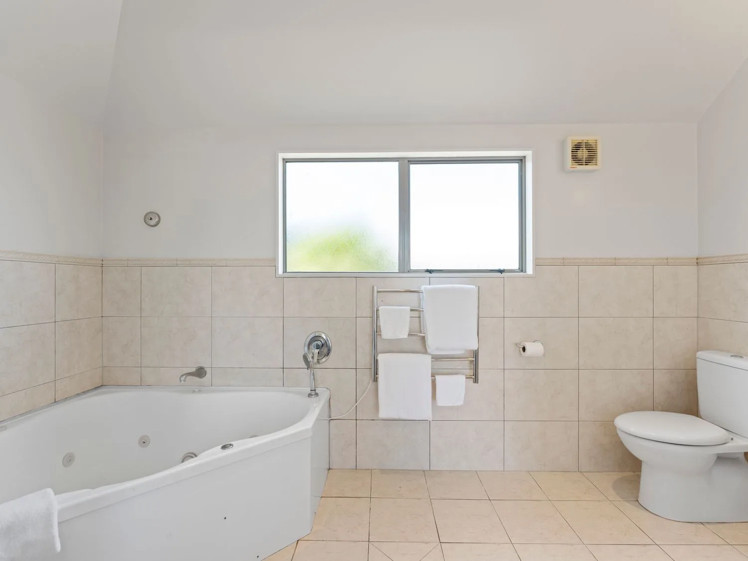 A bathroom with a corner bathtub toilet window and towel rack with towels at Sun and Swim - Lake Taupo Holiday Home Taupo Central