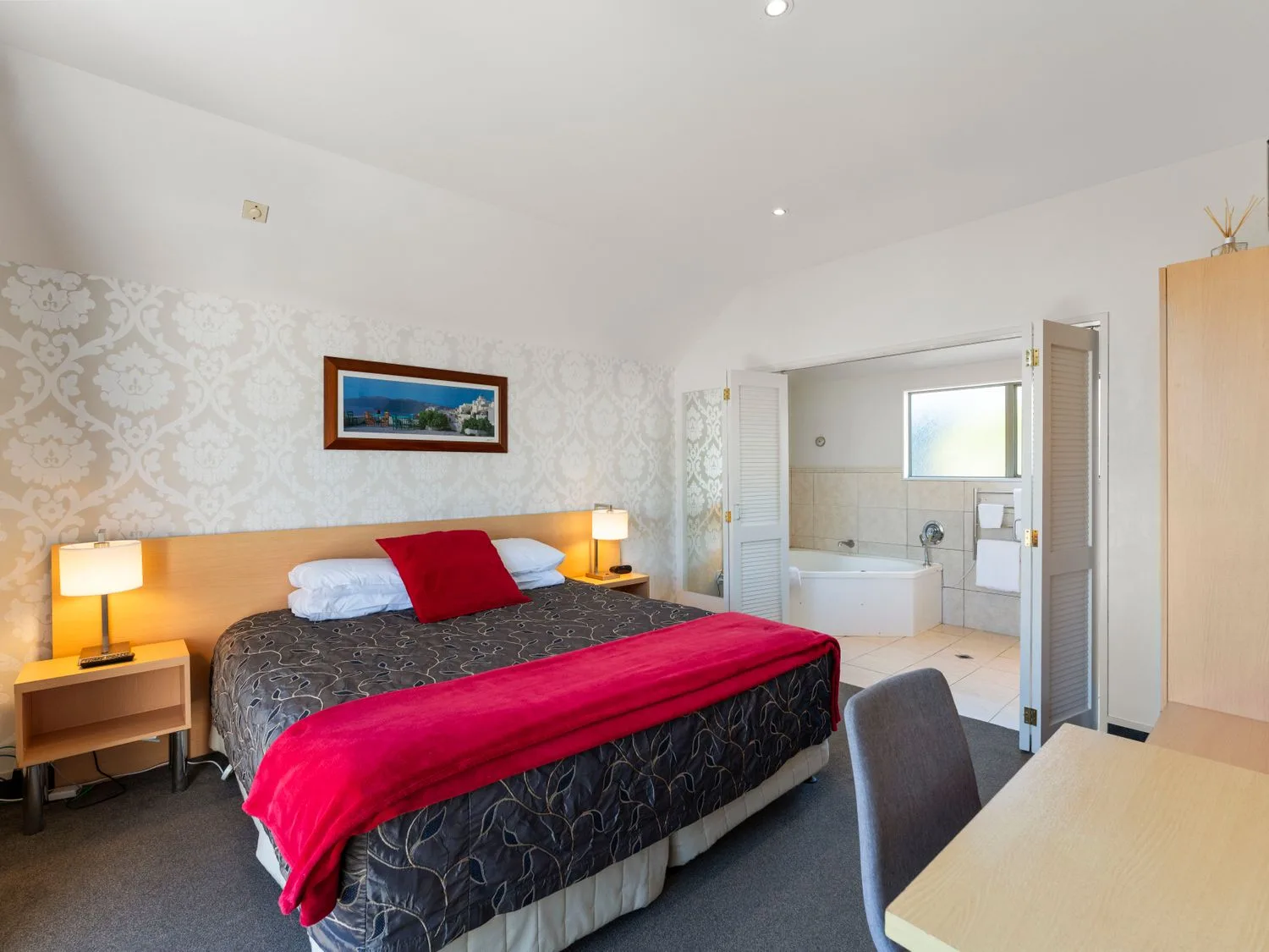A bedroom with a bed covered in dark bedding and a bright red throw beside an open bathroom.