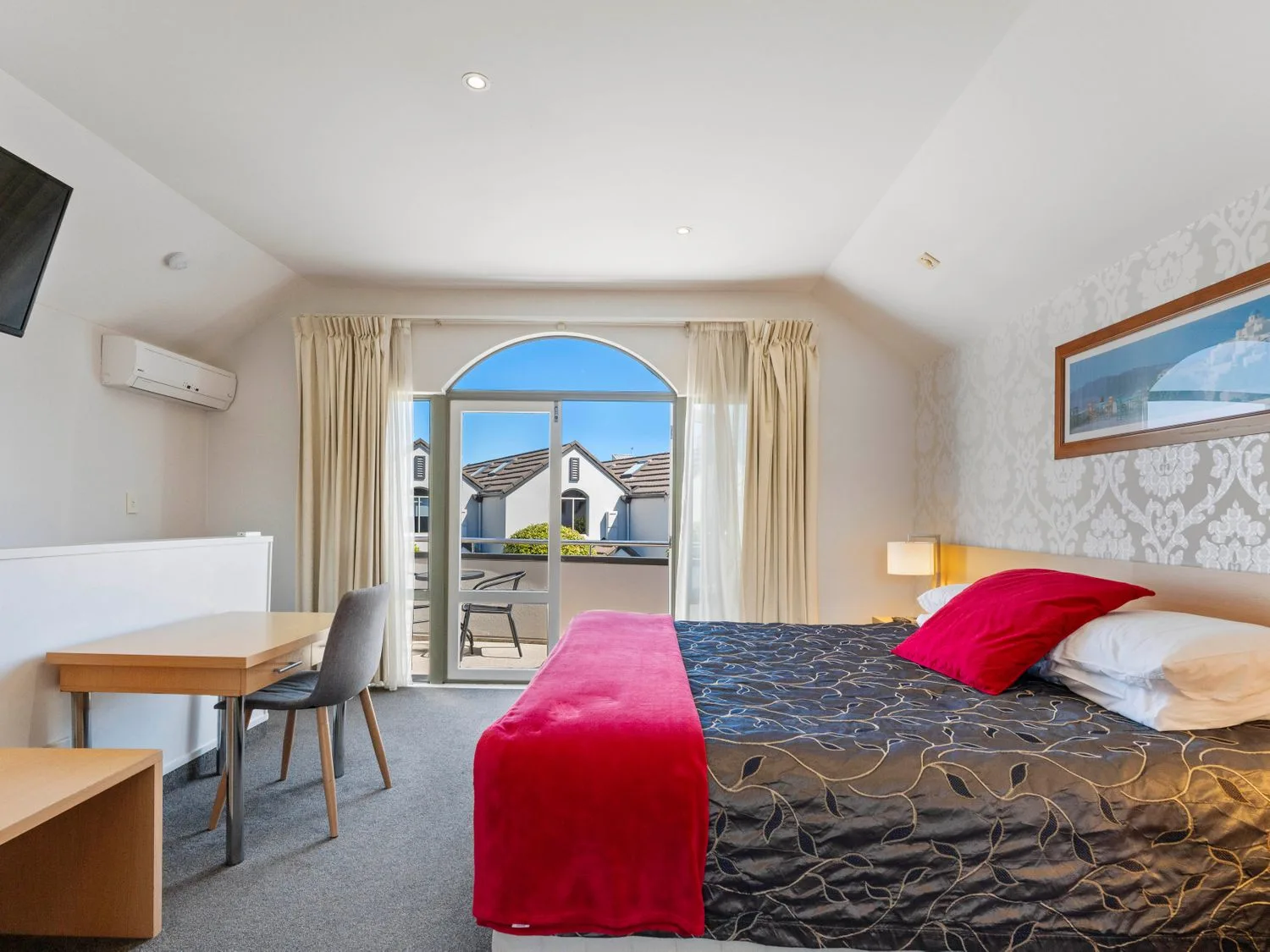 A bedroom with a bed a desk a chair and a balcony at Sun and Swim - Lake Taupo Holiday Home Taupo Central