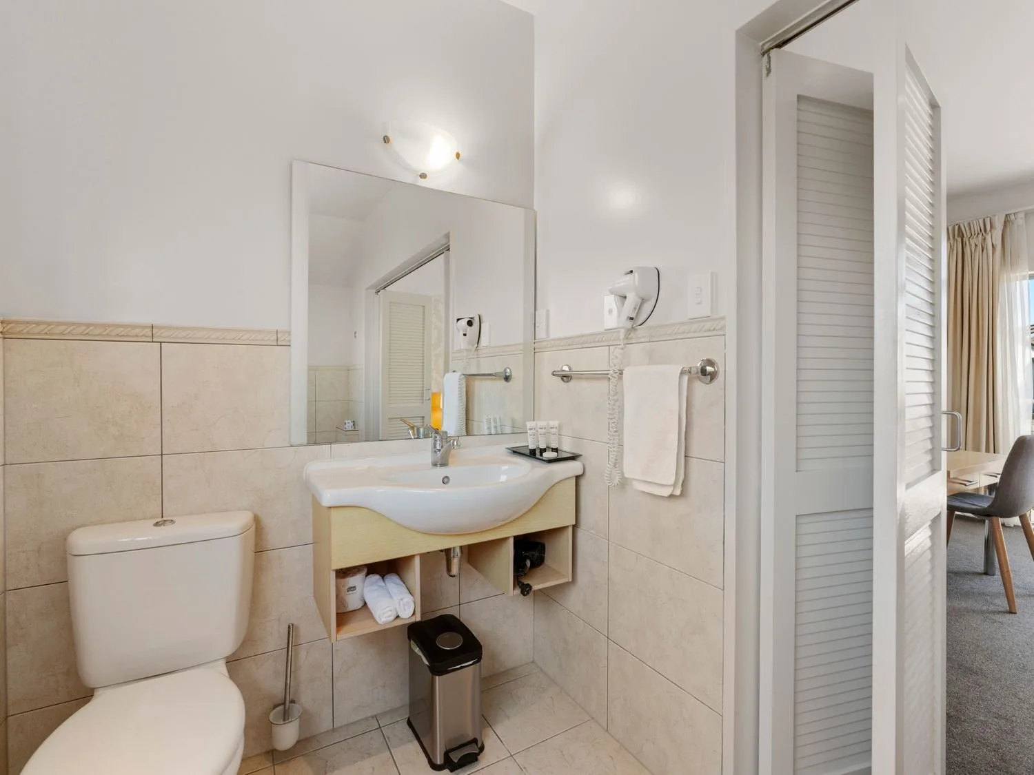 A bathroom with a toilet, a sink with towels and toiletries, a mirror, and a hairdryer at Sun and Swim - Lake Taupo Holiday Home Taupo Central