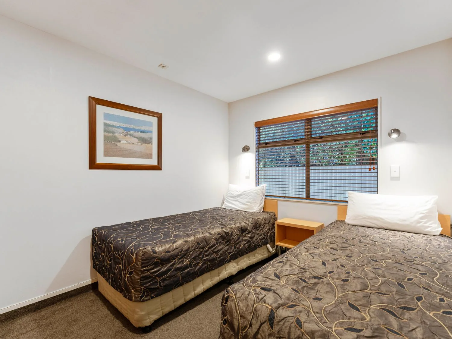 A bedroom with two single beds a wooden nightstand a window with blinds and a framed picture on the wall at Sun and Swim - Lake Taupo Holiday Home in Taupo Central