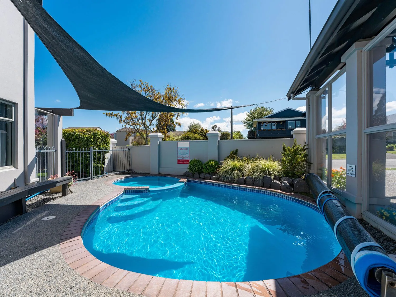 A backyard pool sits beneath a shade sail, with plants along the fence.