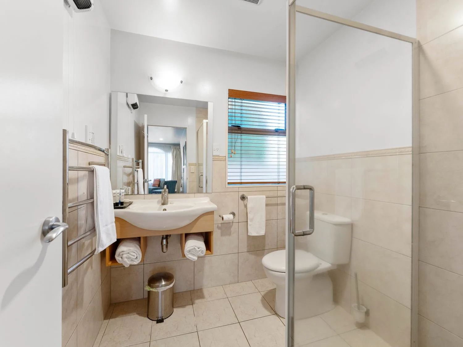 A bathroom with a sink vanity, mirror, toilet, shower, and window with blinds.