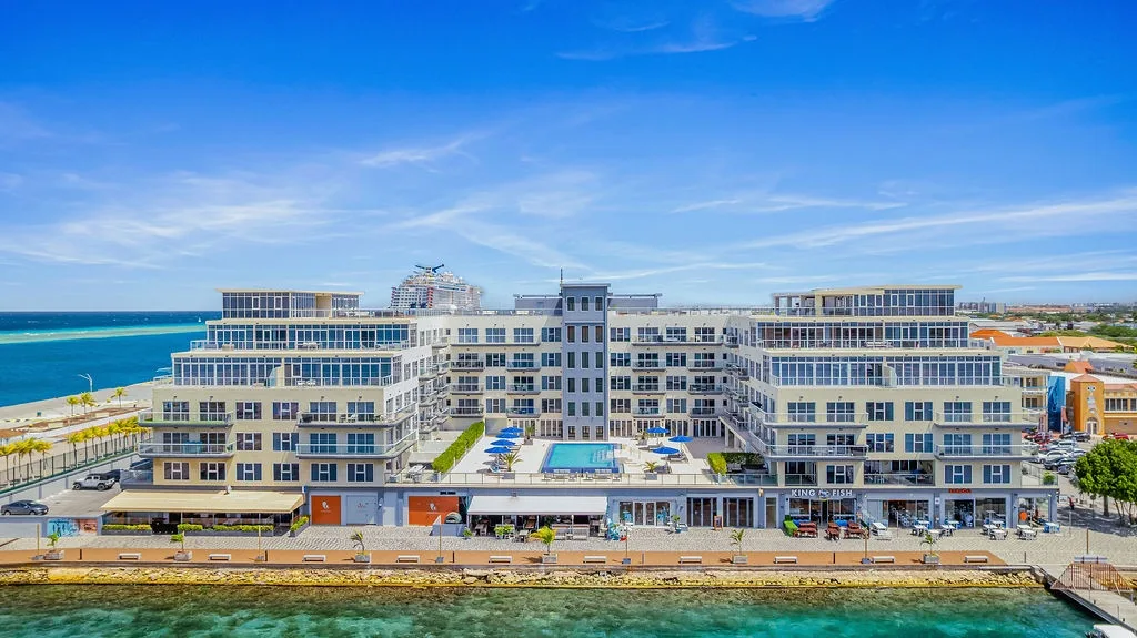 Modern beachfront property with contemporary architecture, positioned directly along turquoise coastal waters with mountain views in the distance and nearby urban amenities.