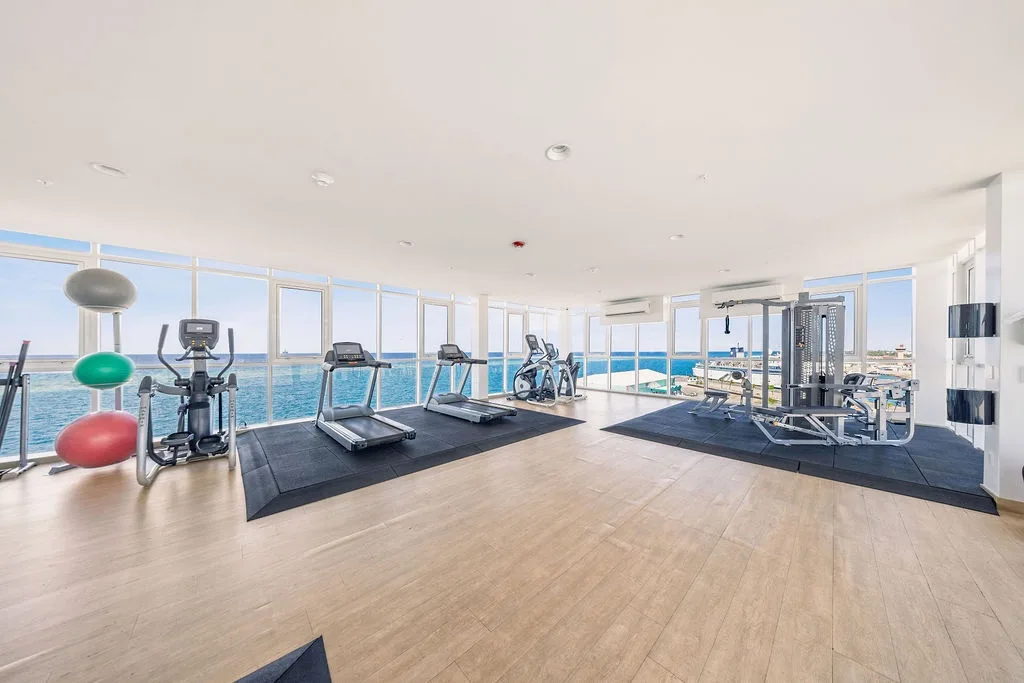 A modern fitness center spans floor-to-ceiling windows overlooking the sparkling sea, where treadmills, bikes, and weights await your waterfront workout.