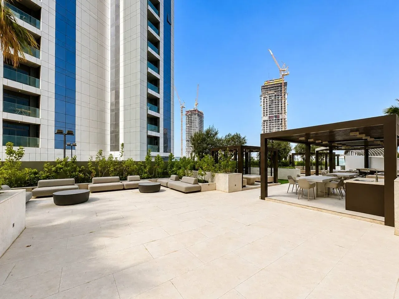 Una terraza exterior con asientos bajo una pérgola sombreada, junto a edificios altos.