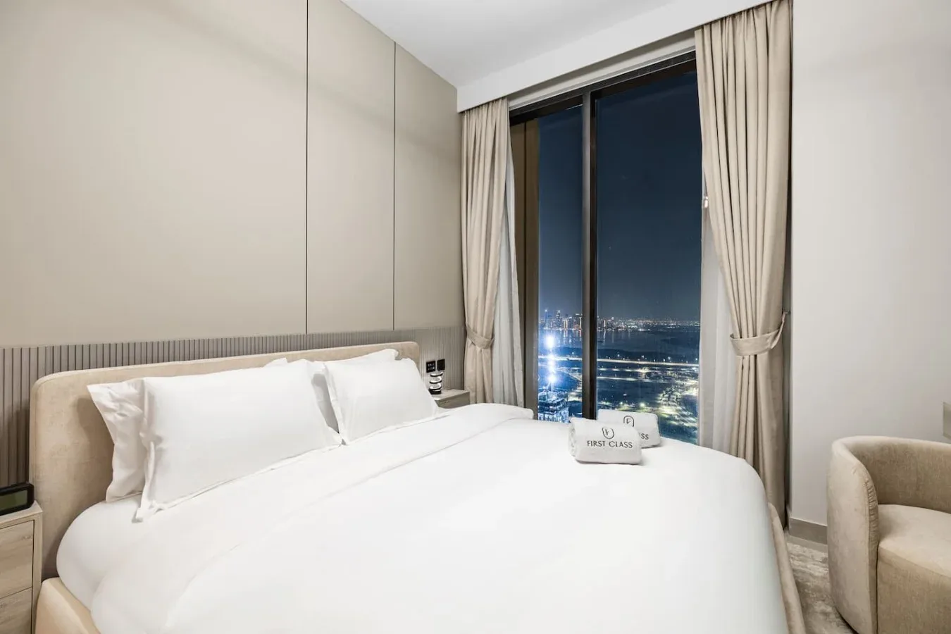 Un dormitorio con una cama tendida junto a una ventana con vistas a las luces de la ciudad.