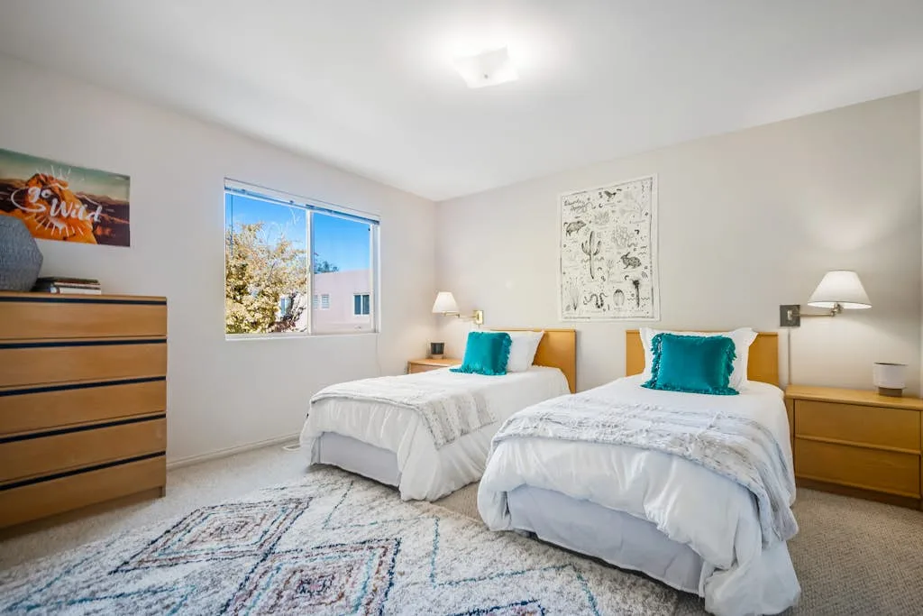 Un dormitorio luminoso con dos camas, cojines turquesa y una ventana.