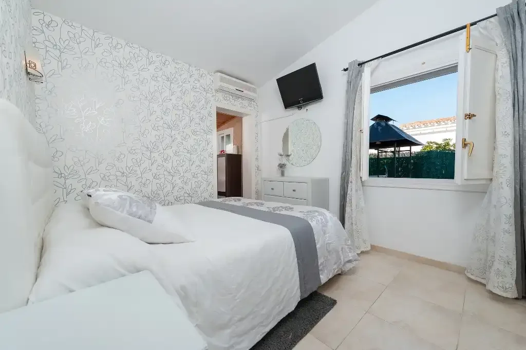 A bright bedroom with white bedding and a window overlooking greenery.