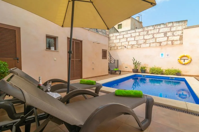 A swimming pool and lounge chairs sit in a sunlit courtyard beneath a large umbrella.