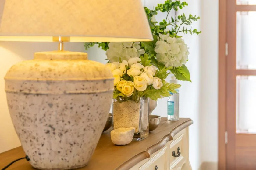 A bedside lamp and flower arrangements sit on a wooden dresser beside a window.