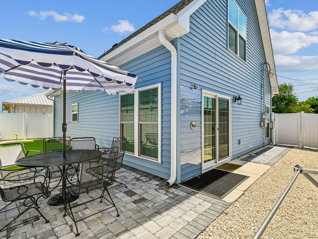 A blue-sided house stands beside a paved patio with outdoor seating and an umbrella.