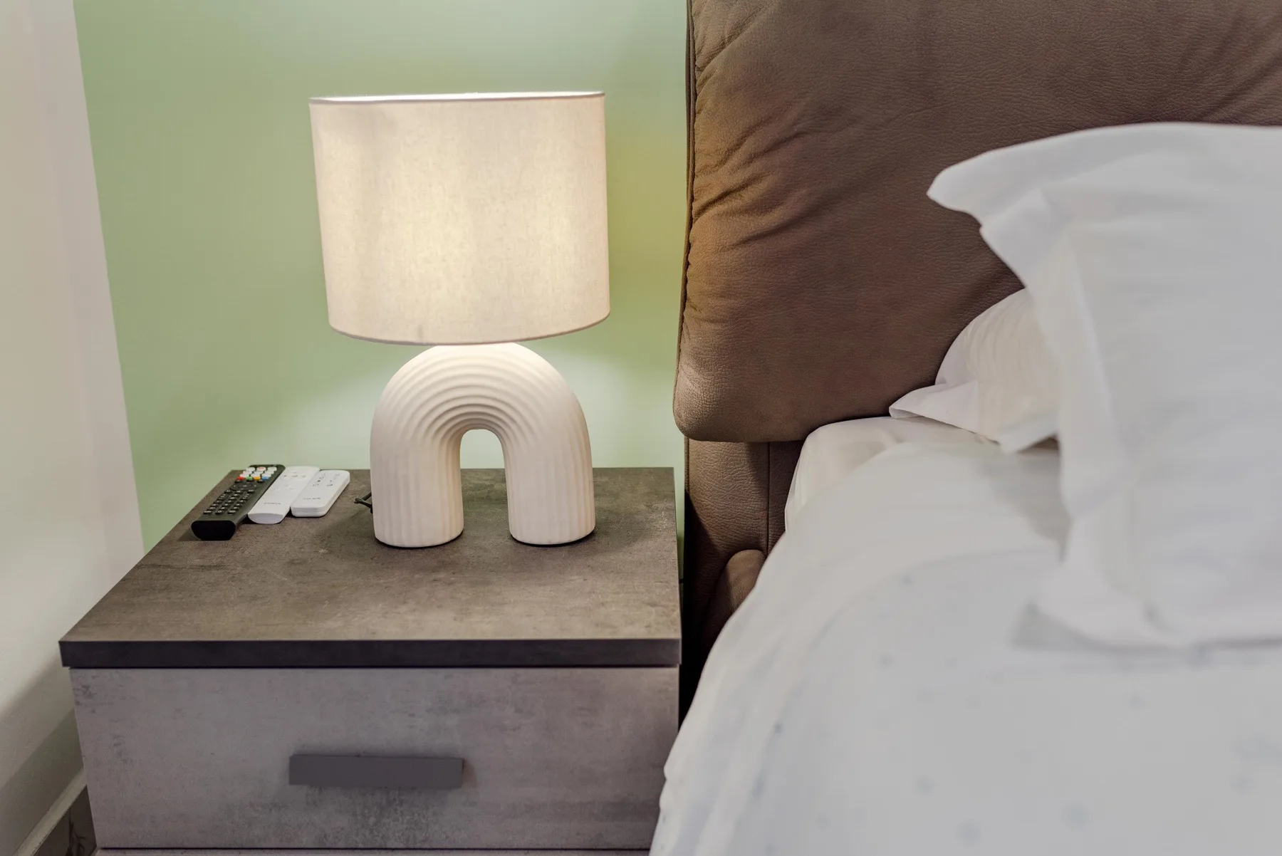 A bedside table with a lit lamp sits beside a bed.