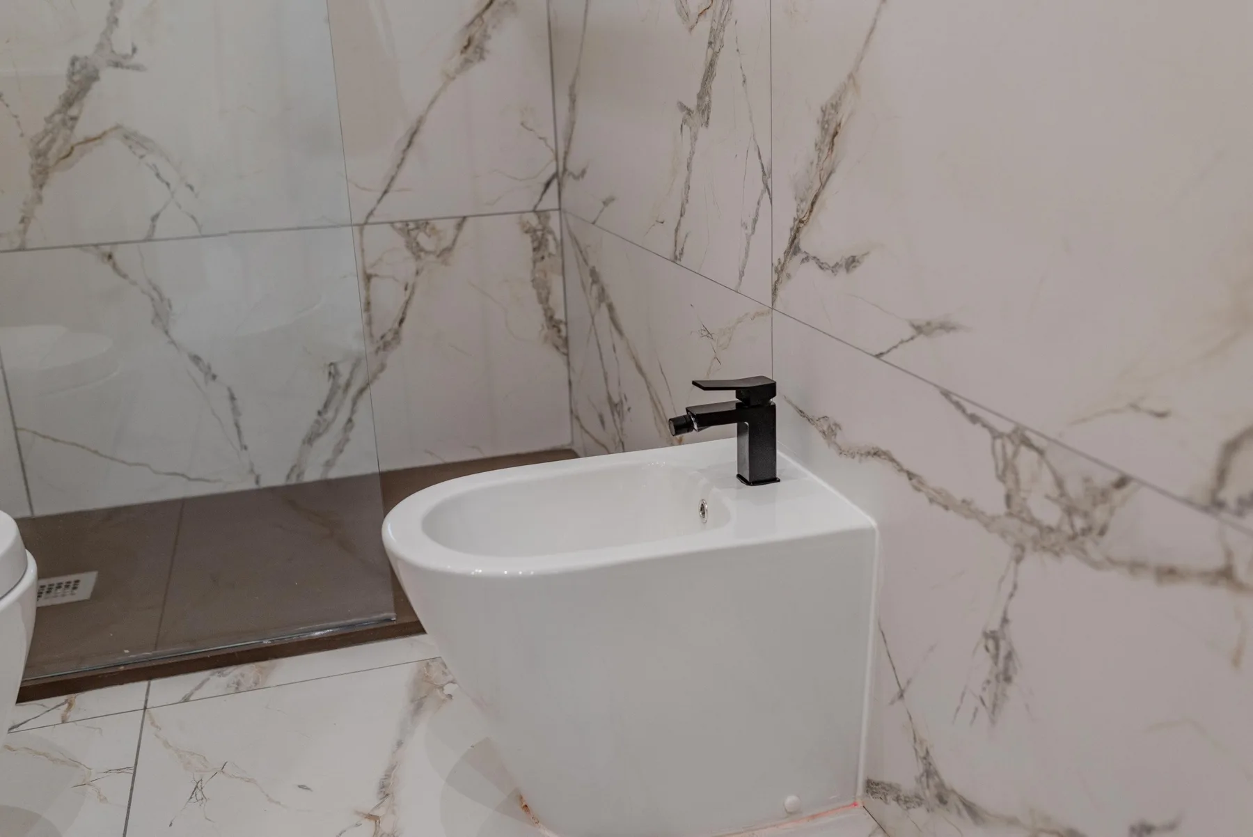 A bathroom with a white bidet and dark faucet against veined tile walls.