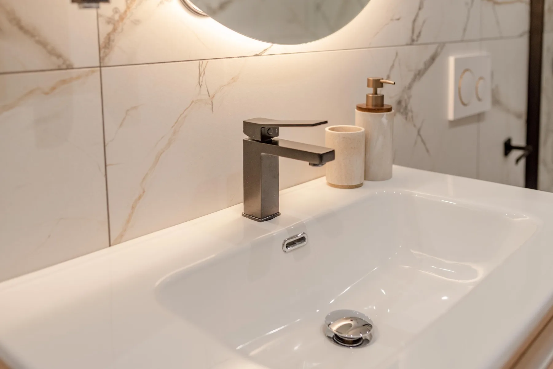 A bathroom sink sits beneath a round mirror, with soap dispensers beside the faucet.