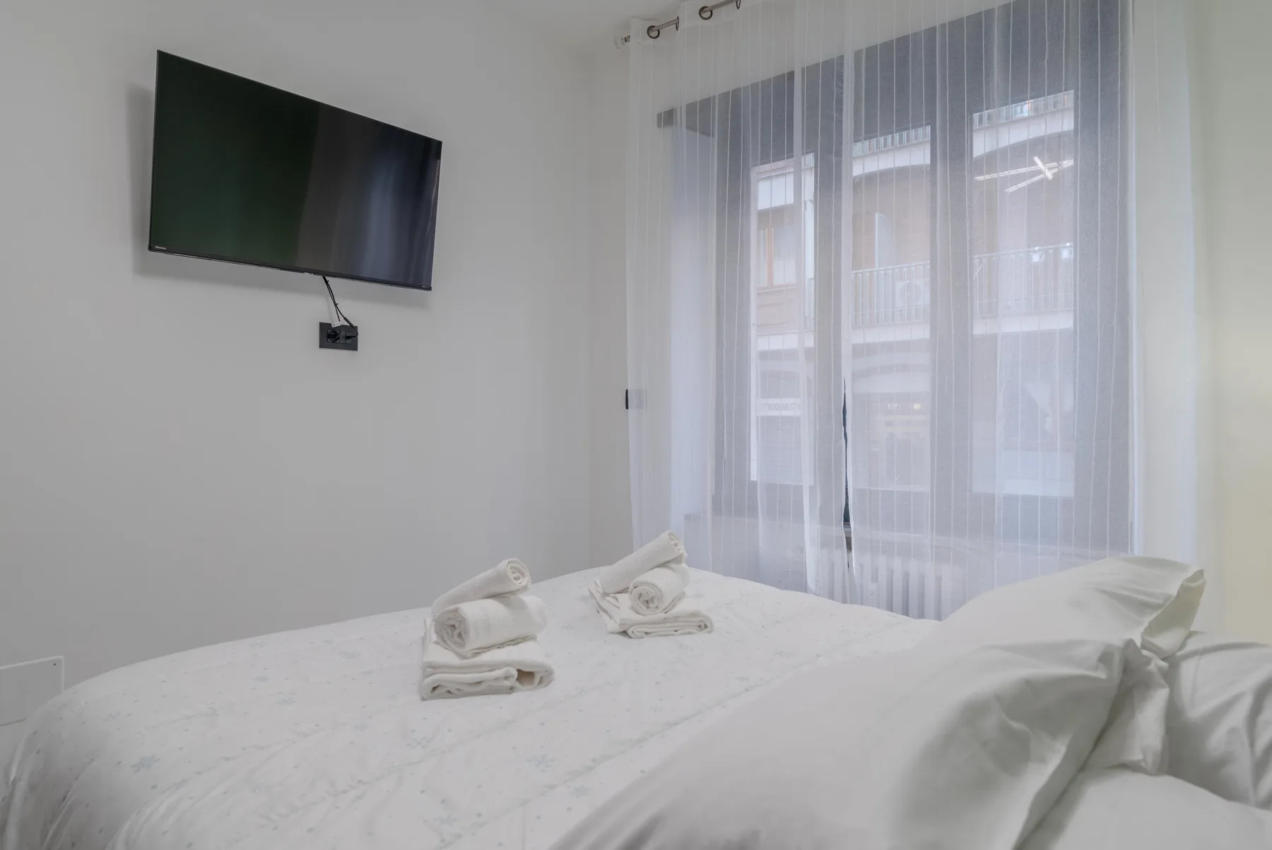A bedroom with a bed, folded towels, and a wall-mounted television.