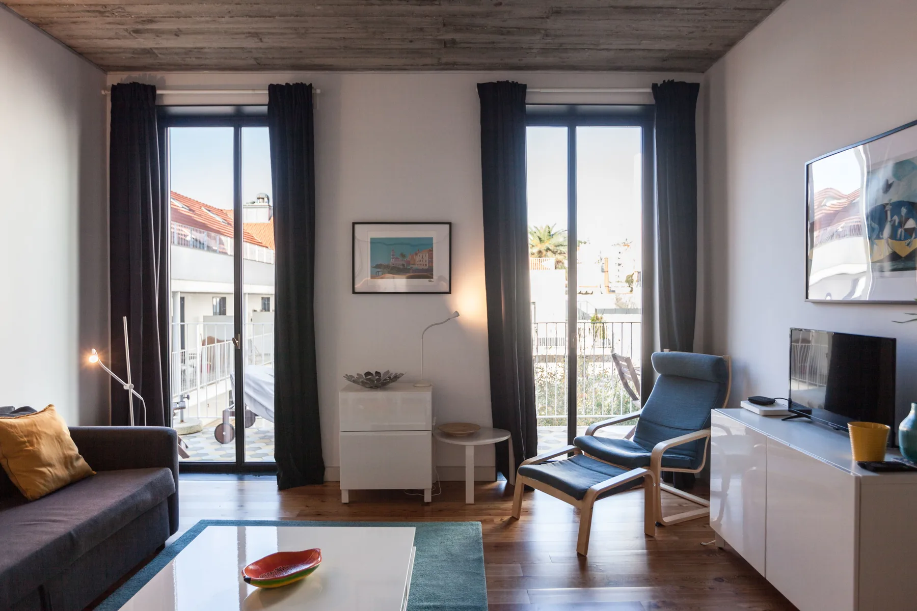 Un salón luminoso con asientos, un televisor y ventanas altas que dan a un balcón.
