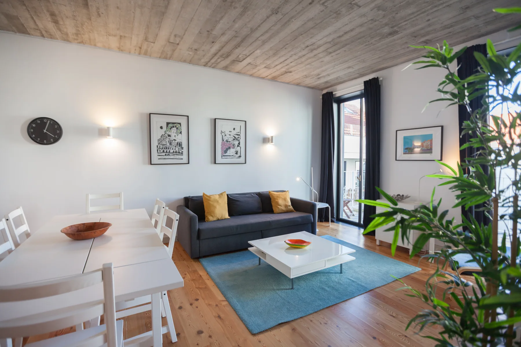 Sala de estar con asientos junto a una mesa de comedor y ventanas altas.