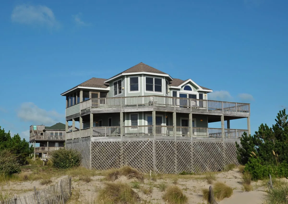 A large coastal house stands on sandy dunes beneath a clear blue sky.
