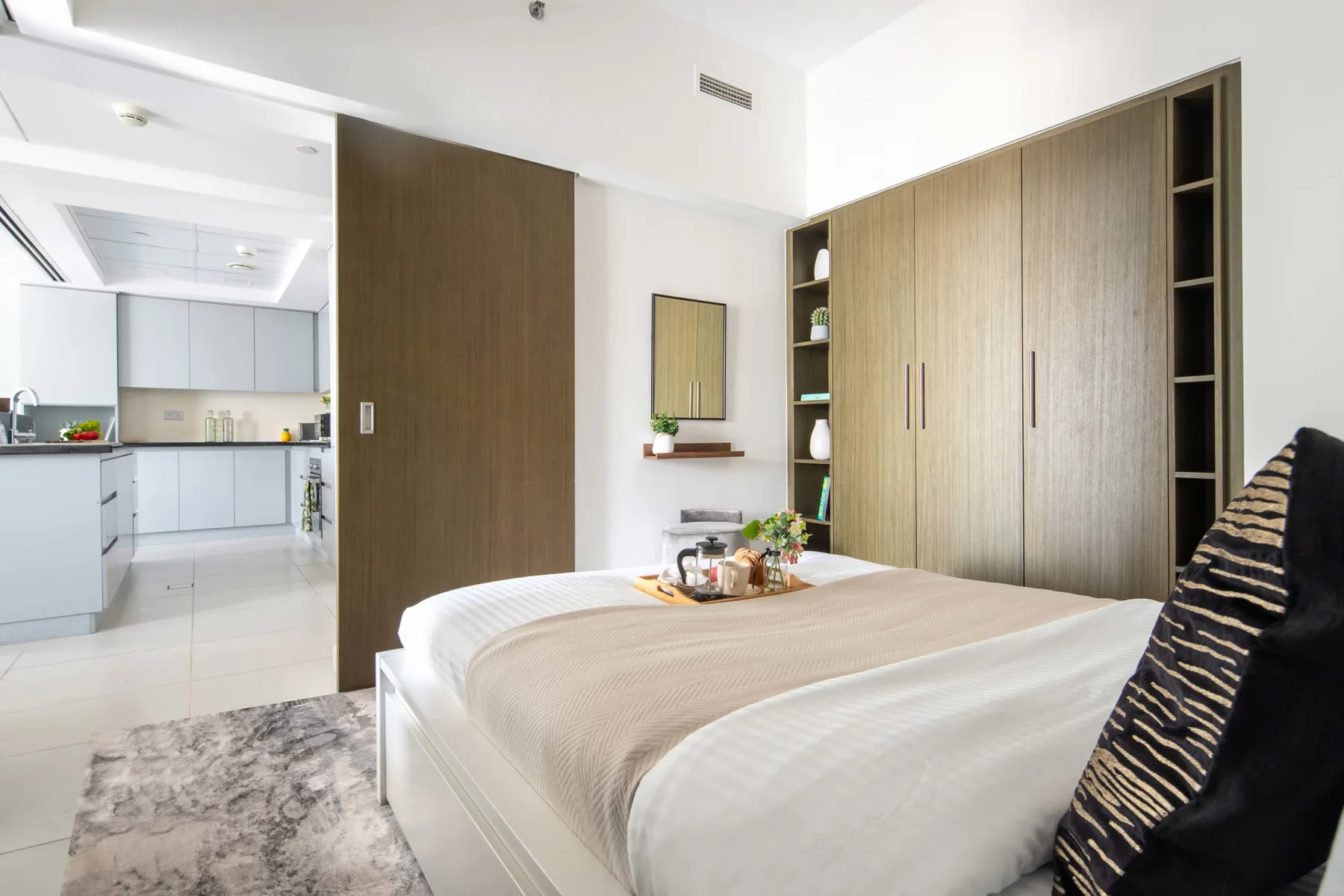 Un dormitorio con una cama tendida y la cocina visible a través de la puerta abierta.