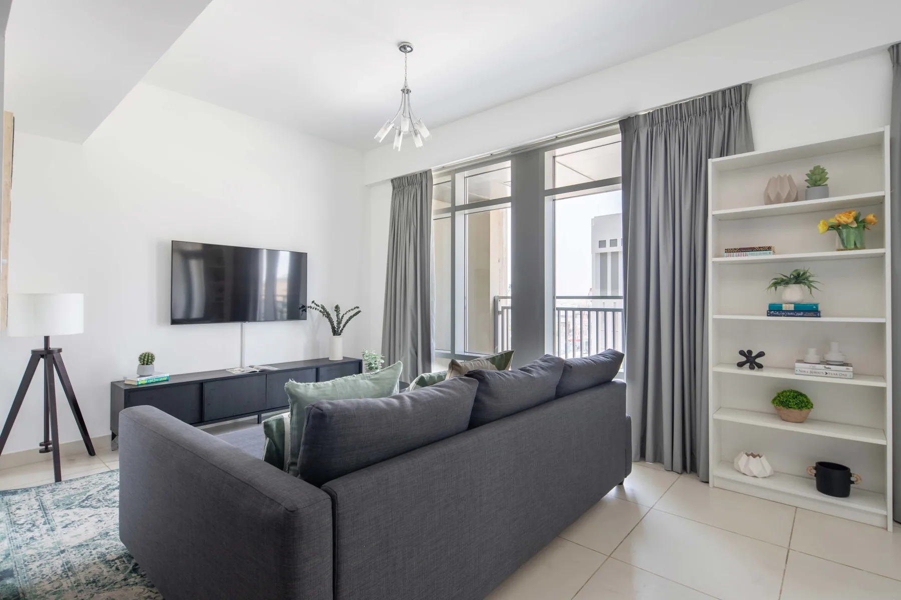 Una sala luminosa con asientos grises, un televisor en la pared y ventanas altas con cortinas.