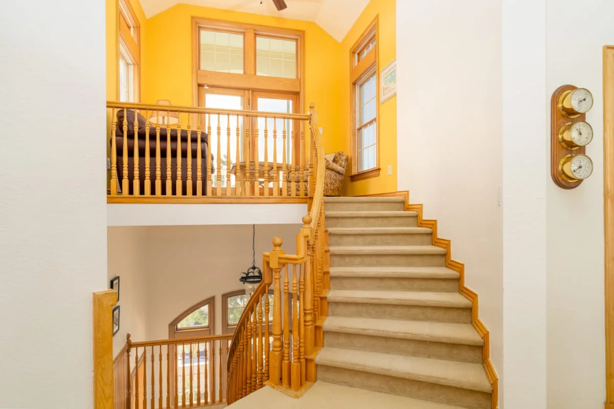 Una escalera con barandillas de madera sube por un interior luminoso de paredes amarillas.