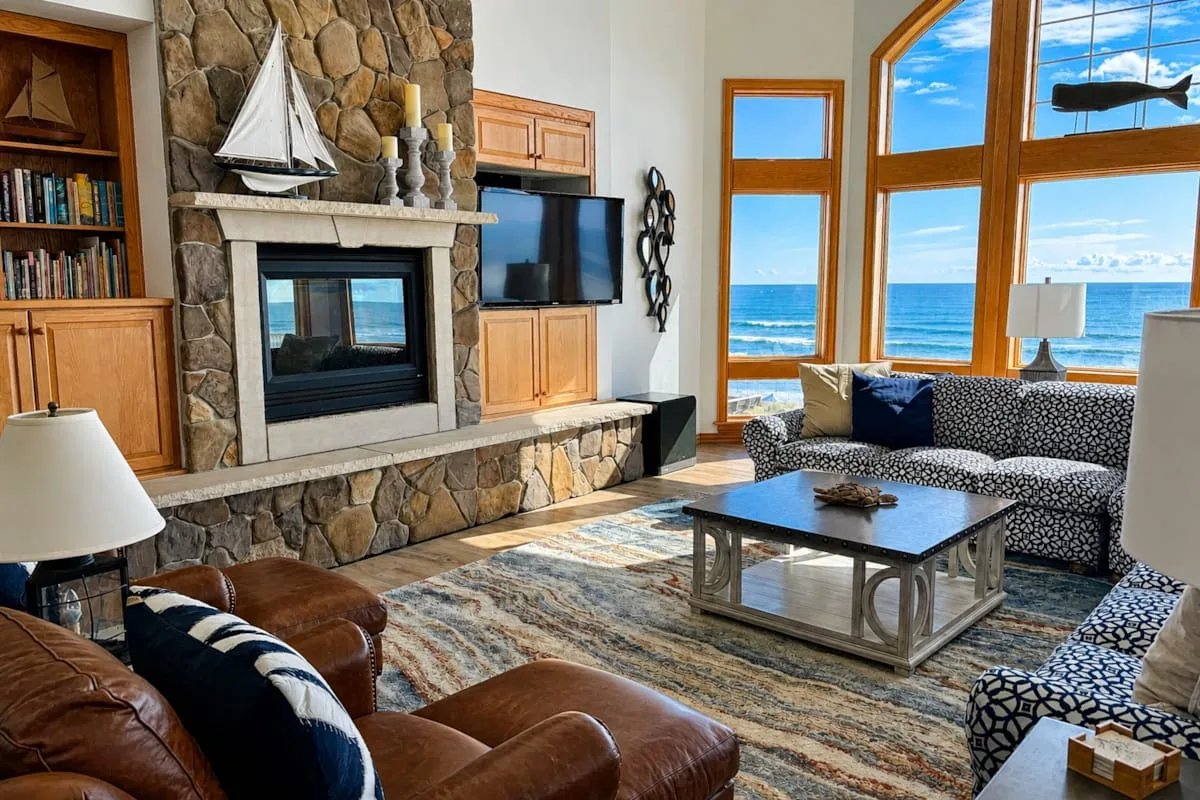 Una sala de estar con chimenea de piedra, asientos y grandes ventanas con vista al océano.