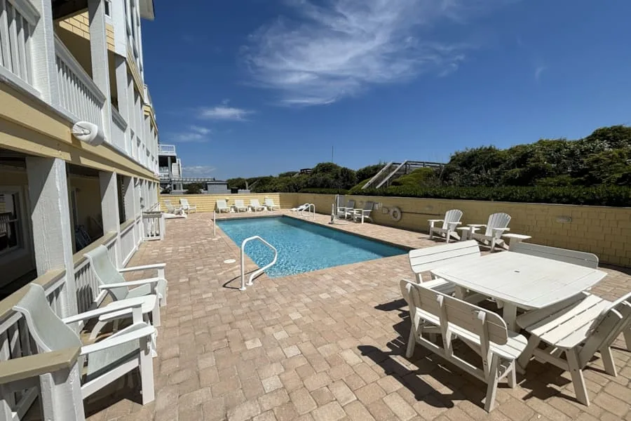 Una piscina al aire libre junto a una terraza pavimentada con asientos y mesas blancas.