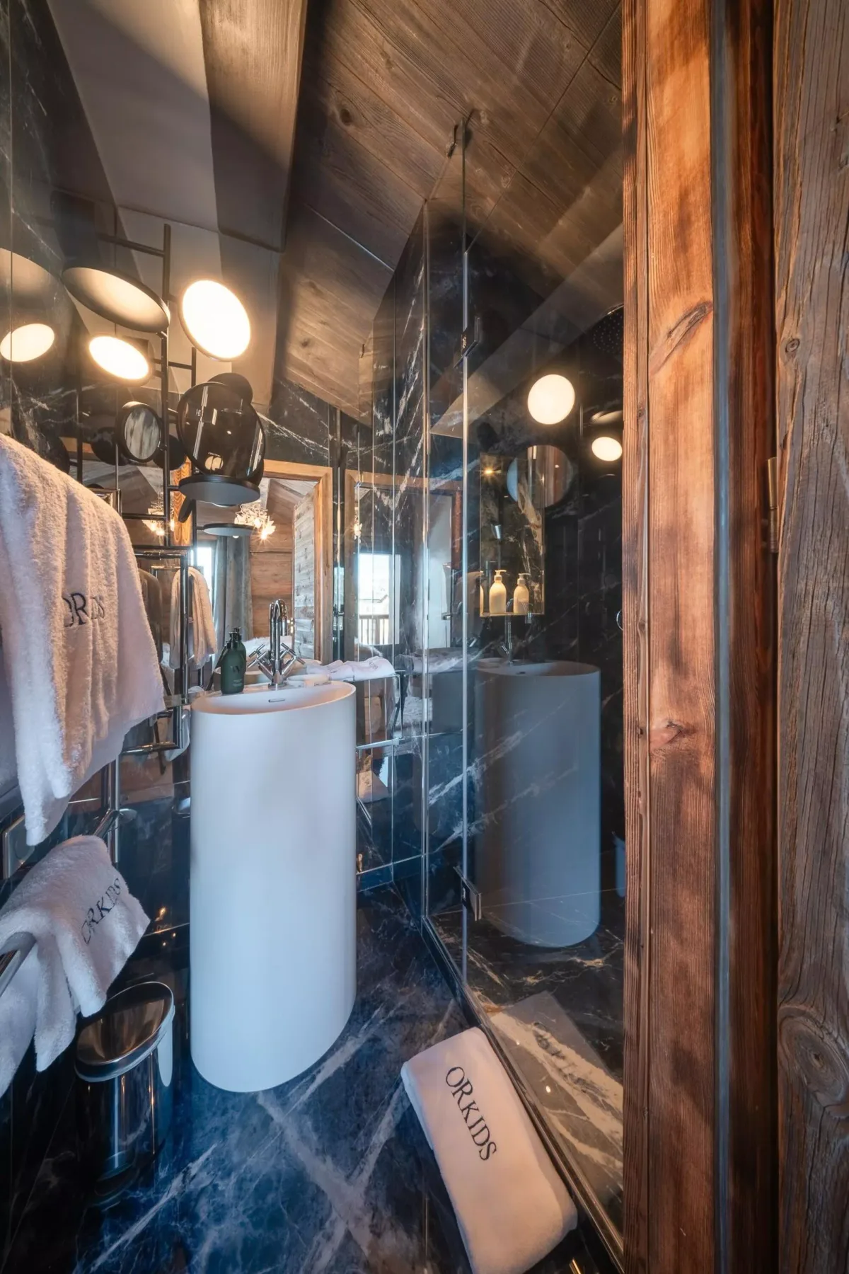 Un baño con un lavabo blanco, espejos y un suelo de piedra oscura.