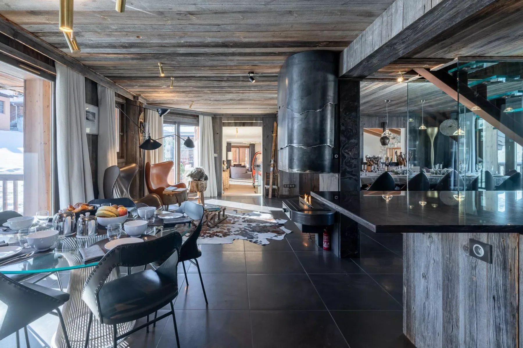 Un comedor moderno se abre a una sala con grandes ventanales y una chimenea oscura.