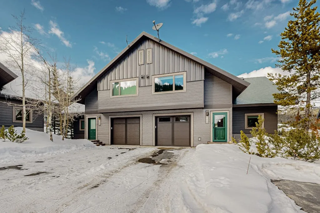 A gray house stands beside a snow-covered driveway under a blue sky.