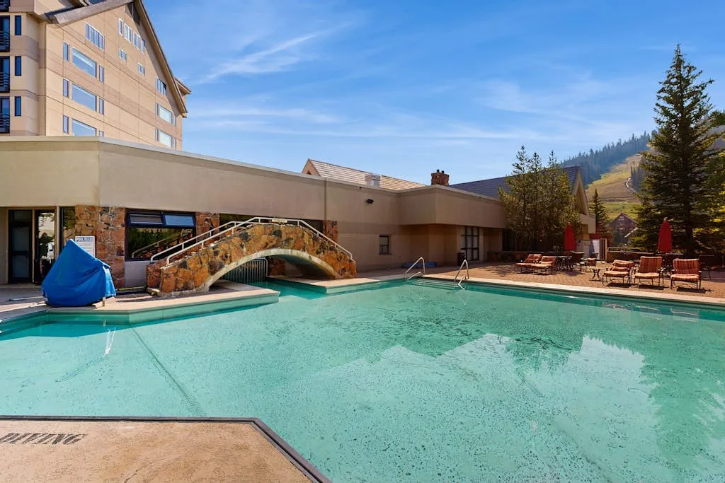 Outdoor swimming pool beside a low building, with a stone bridge over the water.