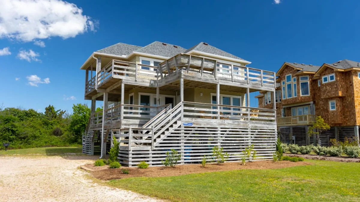 A raised coastal-style house with wraparound decks stands beside a sandy yard.