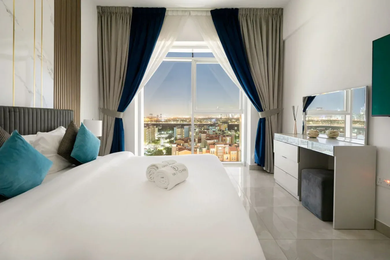 Un dormitorio con una cama bien tendida y una gran ventana con cortinas que da a la ciudad.