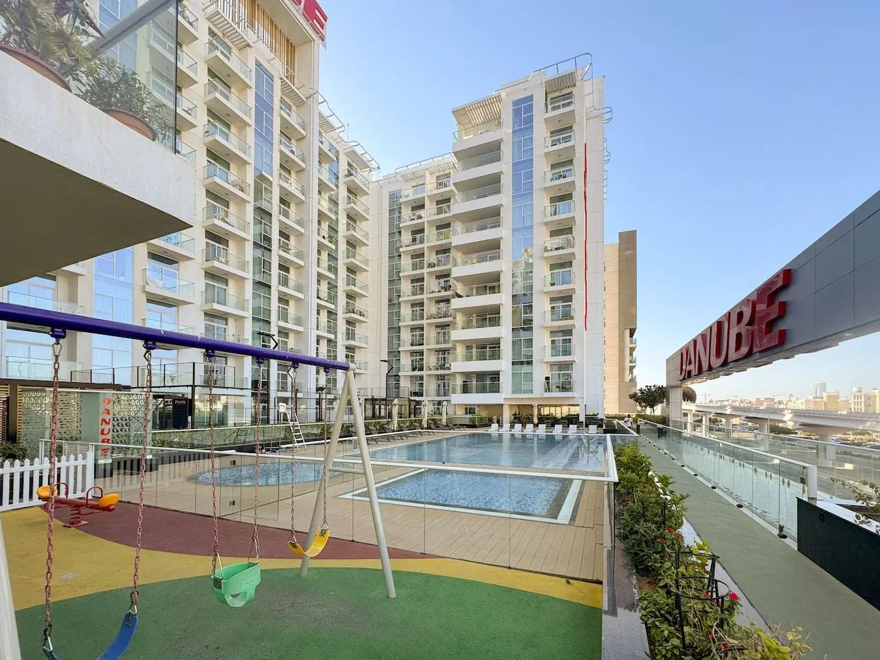 Un moderno complejo de apartamentos rodea una piscina al aire libre y un colorido parque infantil.