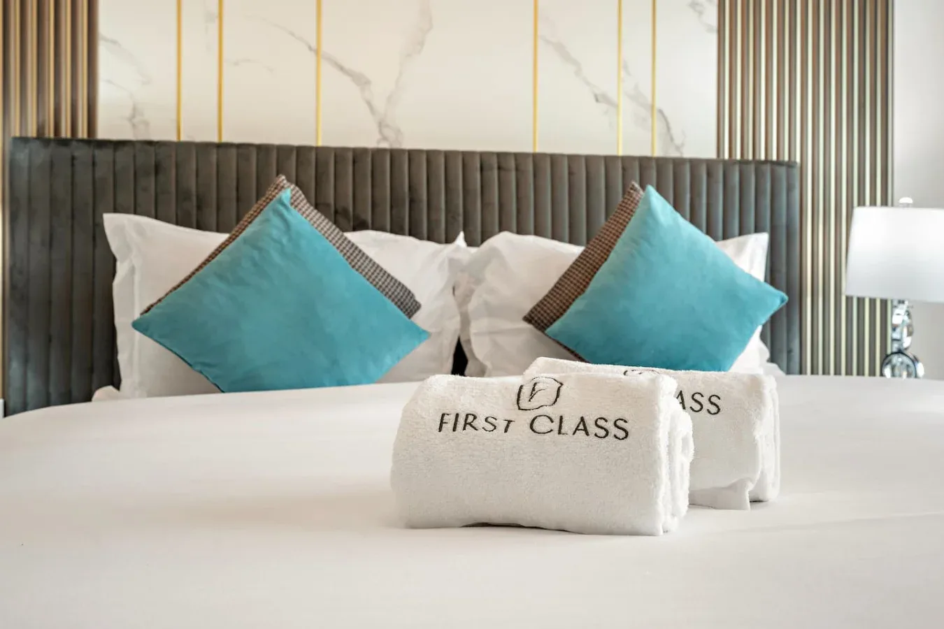 Una cama bien arreglada con almohadas turquesas y toallas blancas dobladas frente a un cabecero oscuro.