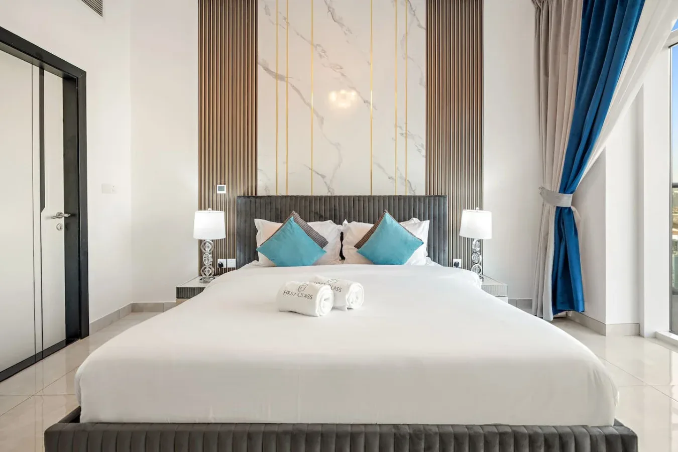 Un dormitorio luminoso con una cama grande, cojines color turquesa y cortinas azules.