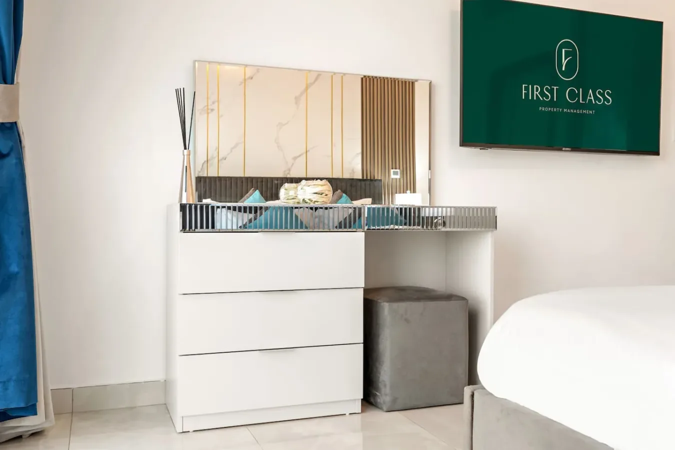Un rincón de dormitorio con una cómoda blanca, un espejo decorativo y el borde de una cama.