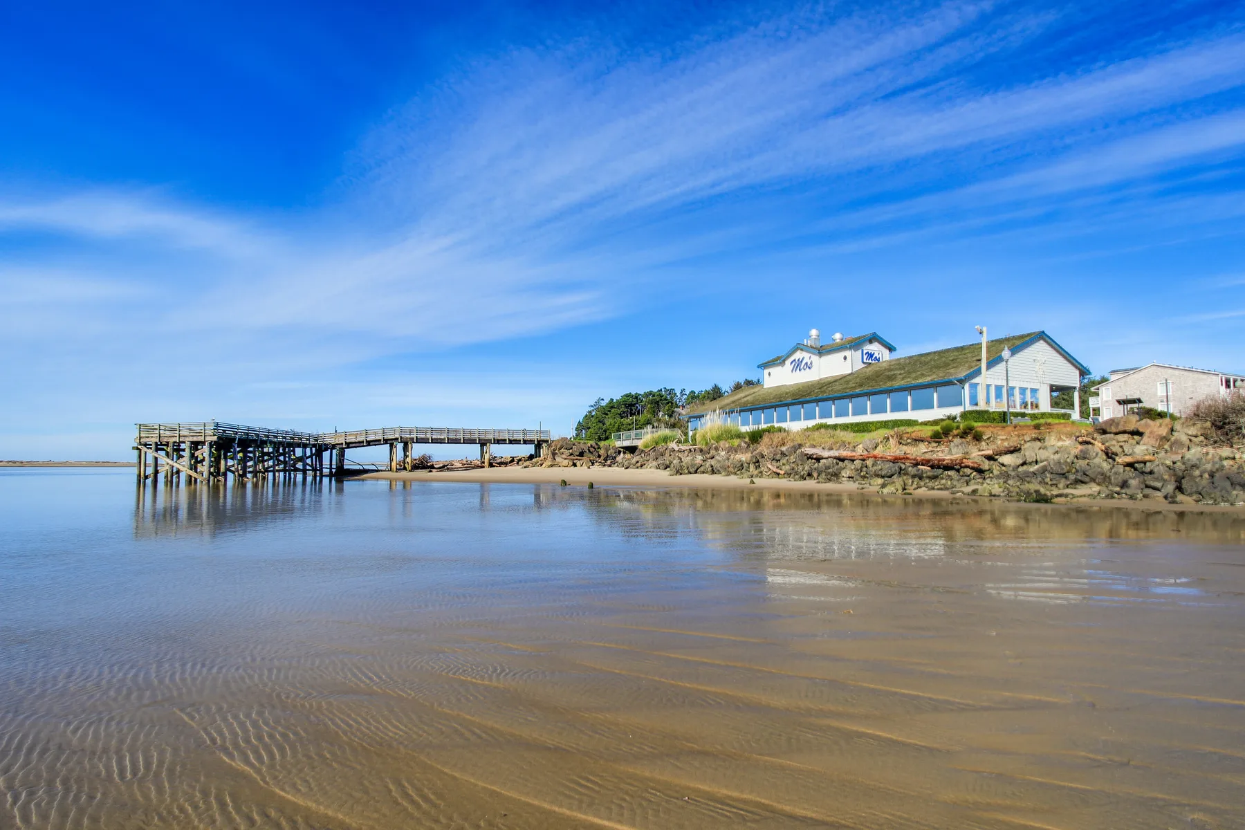 A coastal building stands beside a wooden pier over a calm tidal beach.